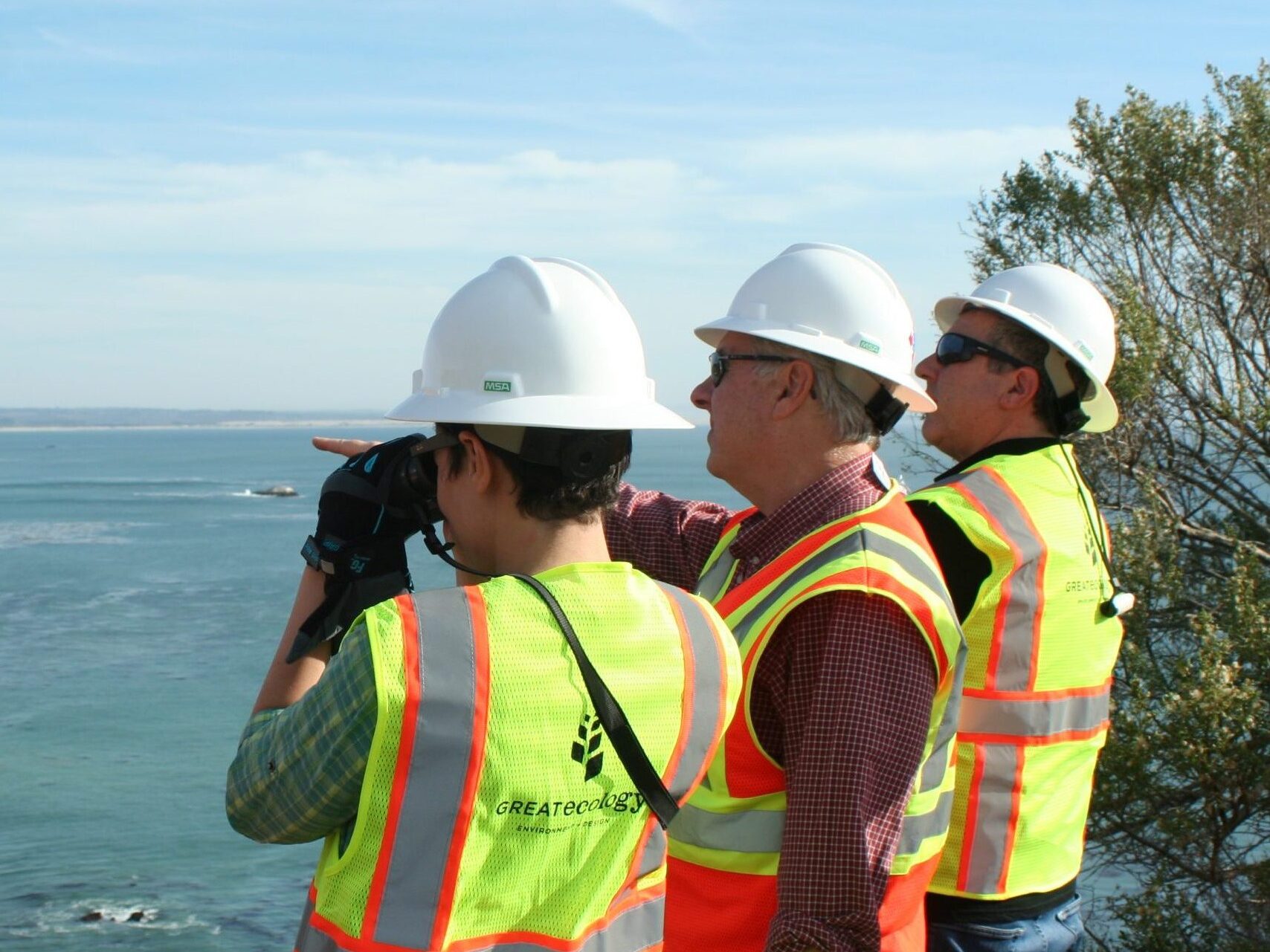 three team members in hard hats and high visibility vests looking out over the coast